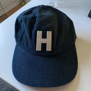 Mörkblå Tommy hilfiger keps  - Snygg mörkblå keps i bomull men ser ut som jeans från Tommy Hilfiger med stor vit H-logga framtill. Justerbart spänne baktill och diskret Hilfiger-detalj. Perfekt accessoar för en avslappnad stil.