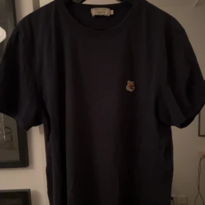 Mörkblå t-shirt Maison Kitsuné - Mörkblå t-shirt från Maison Kitsuné med en broderad rävpatch på bröstet. Klassisk rund hals och korta ärmar. Perfekt för dig som gillar stilrena plagg med en unik detalj. Den är storlek L men passar de flesta med M också