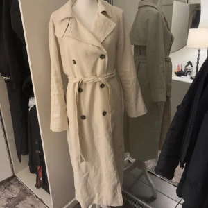 Krämfärgad trenchcoat - Fast pris på 320kr, nypris 699kr  Krämfärgad Trenchcoat – Stilren och Lång 🤍 	•	Längd: 122 cm – jag är 166 cm och den går ner under knäna på mig, perfekt om du letar efter en lång trenchcoat! 	•	Material: Polyester och Viskos – mjuk och bekväm kvalitet  En elegant och klassisk trenchcoat som passar både till vardags och finare tillfällen!