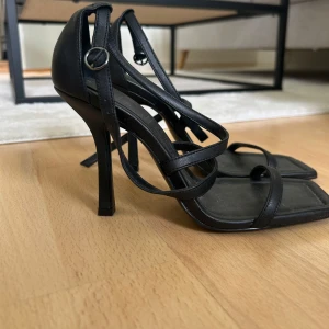 Svarta klackskor - Säljer ett par svarta sandaler med hög klack och smala remmar. Skorna har fyrkantig tå och justerbart spänne vid ankeln. Perfekta för dig som vill ha en stilren och trendig look.