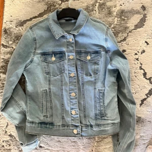 Ljusblå jeansjacka - Säljer en klassisk ljusblå jeansjacka med silverfärgade knappar och två bröstfickor med lock. Jackan har lång ärm, krage och raka sömmar för en tidlös look. Perfekt att slänga över en hoodie eller t-shirt. Använd bara en gång och superfint. Säljer då jag inte tycker jag passar i jeansjackor. 