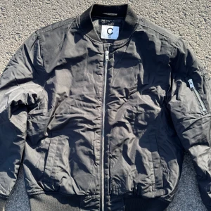 Svart bomberjacka med dragkedja - Säljer en svart bomberjacka från C i storlek 164. Jackan har klassisk bombermodell med ribbade muddar, dragkedja framtill och ficka med dragkedja på ena ärmen. Perfekt för dig som vill ha en stilren och enkel jacka.