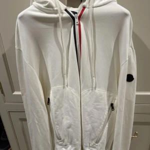 Vit hoodie från Moncler | ev byte - Säljer en stilren vit hoodie från Moncler med hel dragkedja och huva. Jackan har svarta detaljer, fickor med dragkedja och Moncler-logga på ärmen. | ev byte