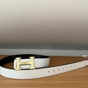 Vändbart bälte med H-spänne - Stilrent vändbart bälte med ikoniskt H-spänne i guld. Ena sidan är vit och den andra svart, vilket gör det enkelt att variera stilen. Bältet  har klassisk design med flera hål för justerbar passform.