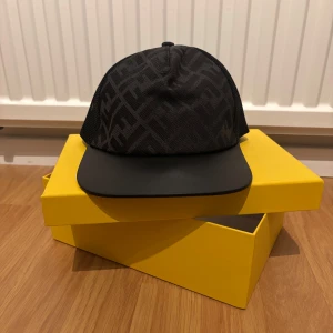 Svart keps från Fendi med mönster - Snygg svart keps från Fendi med diskret mönster och justerbart spänne baktill med gulddetalj. Kepsen är tillverkad i polyester och bomull, har mesh på baksidan för extra ventilation och klassisk böjd skärm.
