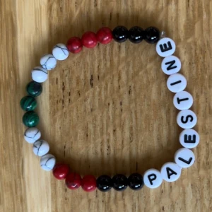 Färgglatt armband med texten PALESTINE - Säljer ett armband med pärlor i grönt, vitt, rött och svart samt vita bokstavspärlor som bildar ordet PALESTINE. Perfekt accessoar för dig som vill visa stöd eller bara gillar färgglada smycken.