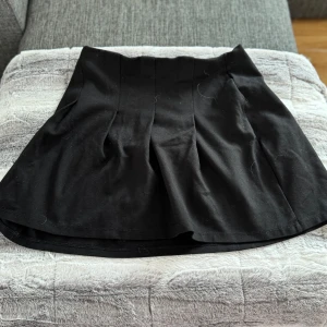 Svart kortkjol med utsvängd passform - Säljer en svart kortkjol med utsvängd siluett och markerade sömmar framtill. Kjolen har en enkel och stilren design som är lätt att matcha med olika toppar. Perfekt för dig som gillar en klassisk look med modern twist.