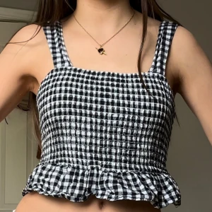 Rutig topp från american eagle - Supersöt croppad topp från American Eagle i svartvitt rutigt mönster. Toppen har breda axelband, smockad framsida och volang nertill. Baksidan har knytning för extra detalj. Perfekt till sommaren och har en fin unik look!!💞