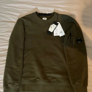 Olivgrön sweatshirt från C.P. Company - Säljer en ny olivgrön sweatshirt från C.P. Company, med rund hals och klassisk passform. Tröjan har en ikonisk linsdetalj på ärmen och ribbade muddar vid ärmslut och nederkant. Perfekt för en stilren och avslappnad look. Köpt på jhonells för 2299 passar xs-s pris in diskuteras