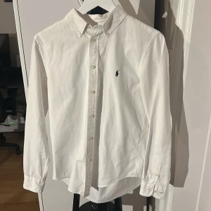 Vit skjorta från Ralph Lauren - Klassisk vit skjorta från Ralph Lauren med broderad logga på bröstet. Skjortan har lång ärm, knappar framtill och button-down krage. Perfekt till stilrena outfits och enkel att matcha med andra plagg.