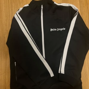 Svart tracksuit från Palm Angels - Säljer en svart track jacket från Palm Angels med vita ränder längs ärmarna och dragkedja framtill. Jackan har hög krage och logga på bröstet. Perfekt för en sportig och trendig look.