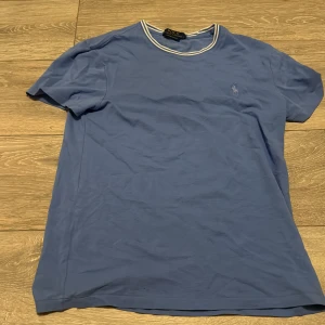 Blå t-shirt från Polo Ralph Lauren - Fräsch t-shirt från Ralph Lauren i ljusblå färg med vit ribbad krage. Klassisk logga på bröstet. Skön och tidlös modell som passar till det mesta. I fint skick! Använt 3 gånger nypris 899