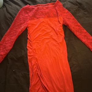 Röd spetsklänning från b.p.c. - Säljer en röd klänning från b.p.c. med långa spetsärmar och blommigt spetsmönster upptill. Klänningen har rynkade detaljer längs sidan och är figurnära med rund halsringning. Perfekt för dig som vill sticka ut med en färgstark look.