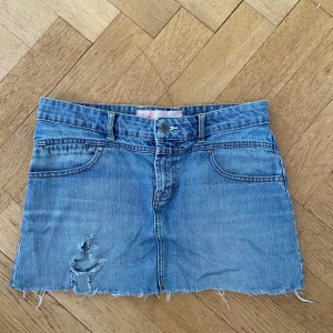 Ljusblå jeanskjol med slitning och hjärtdetaljer - Säljer en ljusblå lågmidjad jeanskjol med rå kant och slitning framtill. Kjolen har två bakfickor med broderade hjärtan och klassiska fickor fram. Perfekt för dig som gillar en avslappnad och trendig stil.