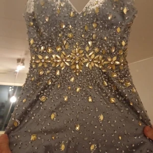 Grå långklänning med guldiga paljetter och stenar - Säljer en riktigt maffig grå långklänning med massor av guldiga paljetter och stenar upptill. Klänningen har v-ringning och markerad midja med blommönster av stenar. Kjolen är i flera lager av tunn tyll som ger en riktigt prinsesskänsla.