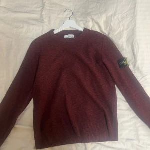 Vinröd tröja från Stone Island - Säljer en vinröd tröja från Stone Island med klassisk rund hals och ribbade muddar. Tröjan har den ikoniska Stone Island-loggan på vänster ärm. Perfekt för dig som gillar stilrena och exklusiva plagg.