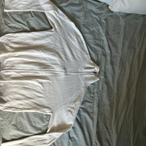 Vit ziptröja i bomull - Säljer en stilren vit ziptröja med lång ärm och dragkedja framtill. Tröjan är i mjuk bomull och passar perfekt för lager-på-lager. Enkel och clean design utan synliga loggor eller mönster.