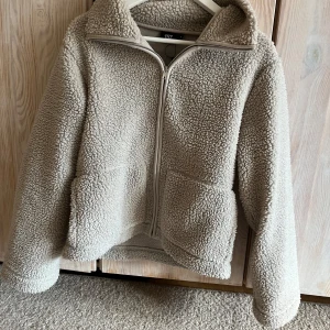 Beige teddyjacka från 157 - Mysig beige teddyjacka från 157 i storlek S. Jackan har dragkedja framtill, två stora fickor och hög krage. Perfekt för kyliga dagar när du vill ha något både snyggt och bekvämt.