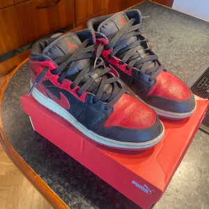 Nike Air Jordan 1 Mid svart/röd - Säljer ett par Nike Air Jordan 1 Mid i svart och rött skinn med klassisk röd swoosh och Air Jordan-logga på sidan. Skorna har svart snörning och platt sula. Perfekta för dig som gillar streetwear och sneakerskultur. De tvättas innnan de skickas 