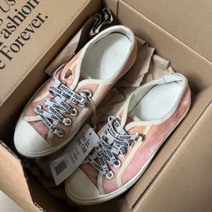 Rosa sneakers från Christian Dior - Säljer ett par exklusiva rosa sneakers från Christian Dior med vita och rosa detaljer. Skorna har vita snören med Dior-tryck, guldiga öljetter och en vit platt sula. Perfekta för dig som vill sticka ut med lyxiga och trendiga skor. De är väl använda och har lite små defekter där av prisetde är köpta på vestiaire collective