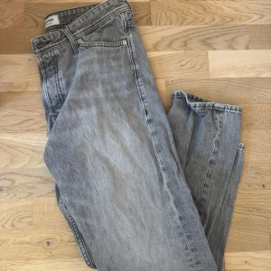 Grå jeans från Jack & Jones - Säljer ett par grå jeans från Jack & Jones med loose fit och klassisk femficksdesign. Jeansen har normal midja och raka ben, perfekt för en avslappnad stil. Färgen är ljusgrå med lätt tvättad look och de stängs med knapp och dragkedja.