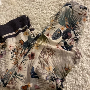 Blommiga byxor med fjärilar från H&M - Säljer ett par tunna byxor från H&M med tropiskt blommigt mönster och fjärilar i olika färger. Byxorna har en ljus bas och inslag av grönt, orange och lila. Perfekta för dig som gillar färg och mönster!