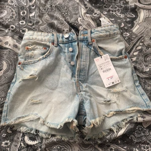 Ljusblå slitna jeansshorts från Zara - Ljusblå jeansshorts med slitna detaljer och fransig kant från Zara. Klassisk femficksmodell med knappar i gylfen och hög midja. Perfekta för en avslappnad stil.