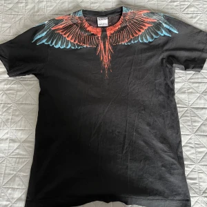 Svart t-shirt med vingmönster från Marcelo Burlon - Säljer en svart t-shirt från Marcelo Burlon County of Milan med ikoniskt vingmönster i rött och blått över axlar och halsringning. T-shirten har rund hals och korta ärmar. Perfekt statement-plagg för dig som gillar streetwear.
