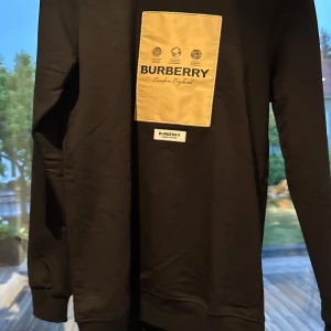 Svart Burberry sweatshirt med beige patch - Svart sweatshirt från Burberry med stor beige patch och tydlig logga på bröstet. Tröjan har rund hals, långa ärmar och en clean, stilren look. Perfekt för dig som gillar exklusiva streetwear-vibbar och vill sticka ut med klassisk design.