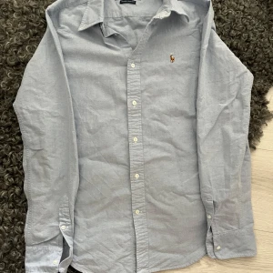 Ljusgrå skjorta från Ralph Lauren - Snygg ljusgrå skjorta från Ralph Lauren i super slim fit-modell. Klassisk design med knappar hela vägen, lång ärm och ikonisk broderad logga på bröstet. Perfekt för dig som gillar stilrena och tidlösa plagg. Skjortan är i mjuk bomull och passar till många olika looks.