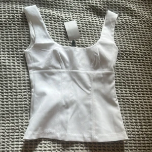Vit korsettopp från H&M - Snygg vit korsettopp från H&M med breda axelband och figurnära passform. Toppen har markerade sömmar framtill som ger en stilren och modern look. Perfekt att styla med jeans eller kjol för en trendig outfit.