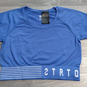 Blå sportig croppad topp H&M - Blå croppad sporttopp från H&M med korta ärmar och bred resår nertill med vita ränder och tryck. Tillverkad i ett stretchigt material som andas, perfekt för träning. Snygg och bekväm passform som sitter på plats under aktivitet.