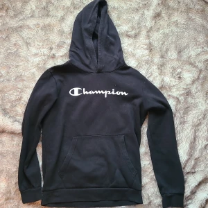 Svart Champion hoodie yrs 13/14 cm.162/167 - Svart hoodie från Champion med vit logga på bröstet. Klassisk känguruficka framtill och huva. Tillverkad i mjuk bomulls- och polyestermix, perfekt för chill dagar eller när du vill ha en enkel streetstyle-look.