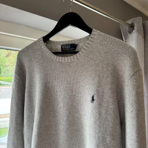 Grå stickad tröja från Polo Ralph Lauren - Snygg grå stickad tröja från Polo Ralph Lauren med klassisk rund halsringning och broderad marinblå logga på bröstet. Tröjan har ribbade muddar vid ärmslut och nederkant, perfekt för lager-på-lager under kyliga dagar.