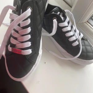 Svarta sneakers från Guess med vita snören - Snygga svarta sneakers från Guess med quiltad ovandel i syntet och textil, vita snören och silverdetaljer med Guess-logga. Skorna har rund tå, platt sula och bling runt snörhålen för extra stilpoäng.
