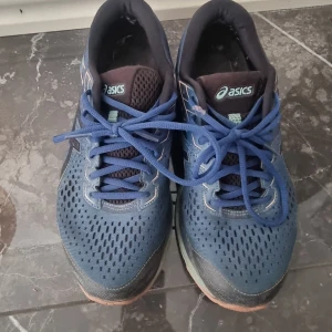 Blå Asics Gel-Cumulus löparskor - Säljer ett par Asics Gel-Cumulus sneakers i blått med svarta och turkosa detaljer. Ovandelen är i ventilerande syntetmaterial och skorna har snörning. Sulan är dämpad med FlyteFoam och har inslag av mintgrönt och korallrosa. Perfekta för löpning eller träning. storlek är 41