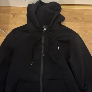Svart Polo Ralph Lauren kofta - Svart hoodie från Polo Ralph Lauren med dragkedja och klassisk vit broderad logga på bröstet. Tröjan har huva med snörning, långa ärmar och känguruficka framtill. Perfekt för en chill och stilren look.