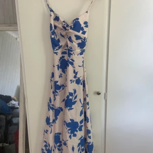 Långklänning med blå blommor och rosett - Superfin långklänning med ljus botten och stora blå blommor. Klänningen har smala axelband, öppen rygg och en snygg rosett bak. Materialet känns lätt och flowy, perfekt för varma dagar. Snygg omlottkänsla framtill och justerbara axelband.
