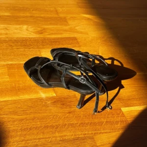Svarta sandaler med klack och remmar - Snygga svarta sandaler med smala remmar och spänne runt ankeln. Skorna har fyrkantig tå och en elegant låg klack. Perfekta för dig som gillar minimalistisk stil och vill ha något stilrent till sommaren.