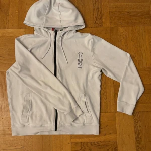 Vit kofta från HUGO med svart tryck - Snygg vit kofta från HUGO med svart grafiskt tryck både fram och stort på ryggen. Tröjan har huva med snörning, dragkedja och två fickor framtill. Perfekt för dig som gillar stilren streetwear med en twist.