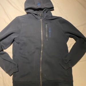 Svart Sail Racing hoodie med dragkedja - Svart hoodie från Sail Racing med hel dragkedja och huva. Snygg blå logga på bröstet och på huvan. Tillverkad i mjuk bomulls- och polyestermix som känns skön mot huden. Perfekt för dig som gillar en stilren och sportig look.