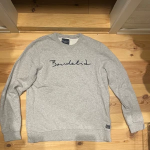 Grå sweatshirt från Bondelid - Grå sweatshirt från Bondelid med broderad logga i mörkblått framtill. Klassisk rund halsringning, långa ärmar och ribbade muddar. Mjuk och skön bomullsmix som passar perfekt till jeans eller joggers. Står XL men sitter som M 
