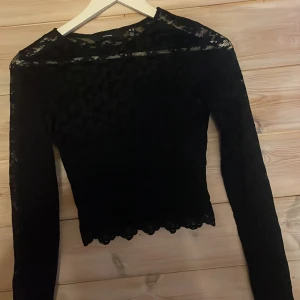 Svart spetsblus från Vero Moda - Säljer en svart långärmad blus från Vero Moda i genombruten spets med fina detaljer. Toppen har rund halsringning och vågig kant. 