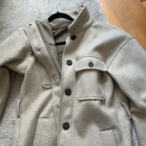 Beige overshirt från ZARA i ullmix XS - Snygg beige overshirt från ZARA i ullmix med stora svarta knappar och flera fickor framtill. Jackan har en rak passform och klassisk krage, perfekt att slänga över en hoodie eller t-shirt. Enkel och stilren vibe som funkar till allt.