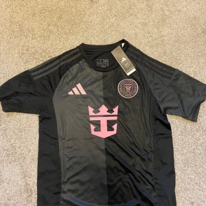 Inter Miami CF svart fotbollströja Adidas - Svart Inter Miami CF fotbollströja från Adidas i slim fit. Tröjan har korta ärmar, klubbmärke och sponsorlogga i rosa framtill samt klassiska Adidas-ränder på axlarna. Tillverkad i lätt och ventilerande material, perfekt för match eller träning.