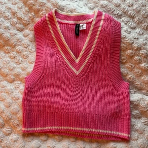 Rosa stickad väst från H&M Divided - Snygg rosa stickad väst från H&M Divided med v-ringning och ribbade kanter. Västen har vita och vita ränder vid halsringning och nederkant, vilket ger en sportig och trendig look. Perfekt att styla över en skjorta eller t-shirt.