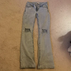 Bootcut jeans i ljusblå denim från Nelly - Bootcut jeans från Nelly i ljusblå denim med slitna hål på knäna och broderade detaljer på bakfickorna. Ordenarie pris 699 kr. Klassisk femficksmodell med låg midja och vida ben. Använda några gånger, men ändå i bra skick förutom nedre delen på benen vid hälen där det bildats fläckar då dom är för långa för mig.