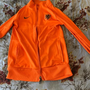 Holländsk orange Nike jacka - Snygg orange jacka från Nike med Nederländernas landslagslogga på bröstet. Jackan har hel dragkedja, ribbade muddar och två fickor framtill. Perfekt för dig som älskar fotboll och vill sticka ut med en sportig stil.