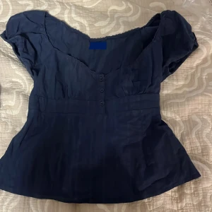 Mörkblå blus med knappar - Snygg mörkblå blus med korta ärmar och v-ringning. Blusen har tre dekorativa knappar framtill och lätt rynkade detaljer vid bysten. Perfekt för dig som gillar en feminin och enkel stil. Aldrig använd