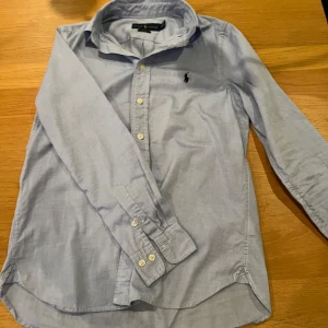 Ljusblå skjorta från Ralph Lauren - Snygg ljusblå skjorta från Ralph Lauren i slim fit-modell. Skjortan har klassisk krage, långa ärmar och knäppning framtill. Ikoniska broderade loggan på bröstet. Perfekt för dig som gillar stilrena och tidlösa plagg.                                                  Passar st xs med